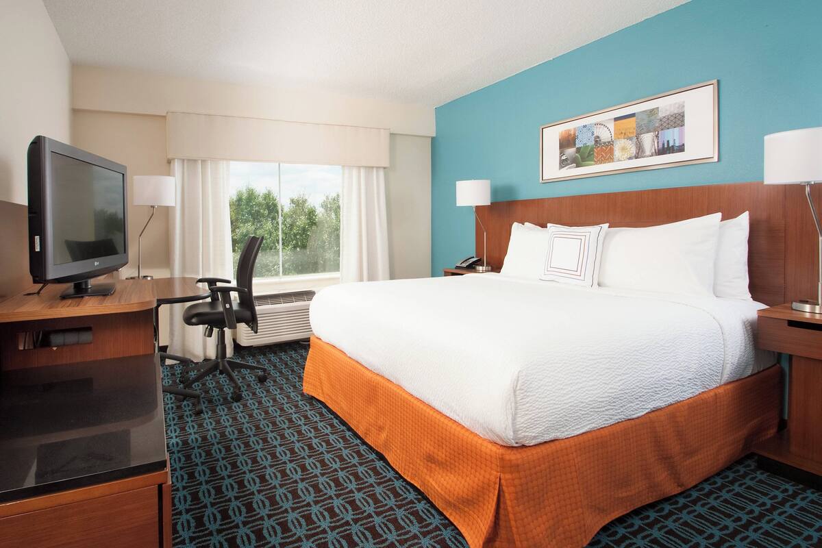 A hotel room with a large bed, a desk with a television, a chair, and a window with curtains.