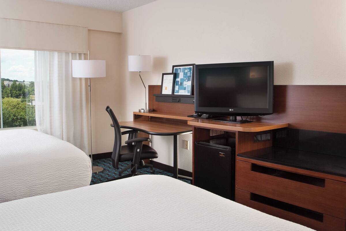 A hotel room with two beds, a desk with a computer and television, a chair, and a lamp.