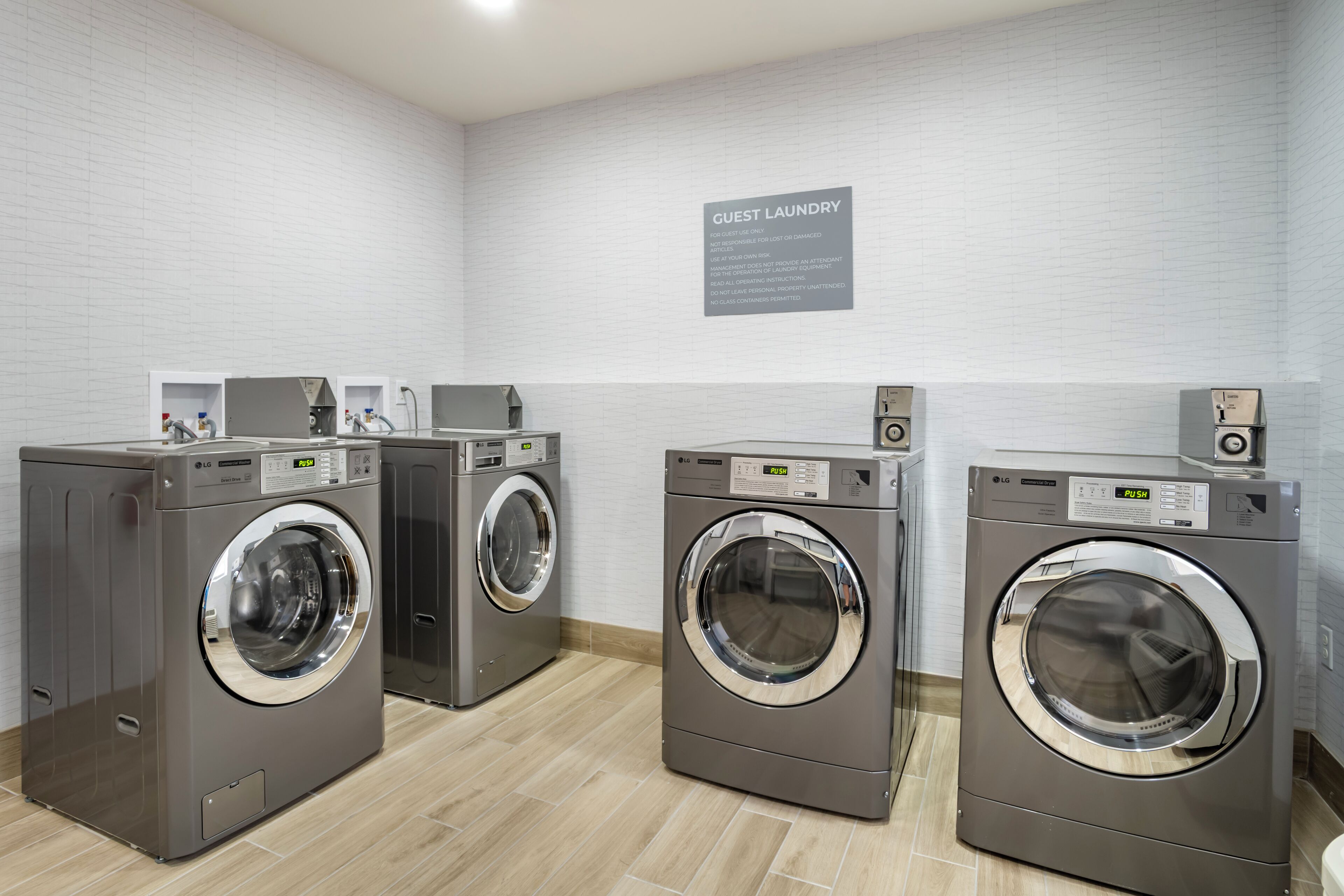 Une laverie équipée de trois machines à laver et de trois séchoirs LG à usage commercial, avec une enseigne indiquant « Laverie pour les clients » et un mur en briques blanches.