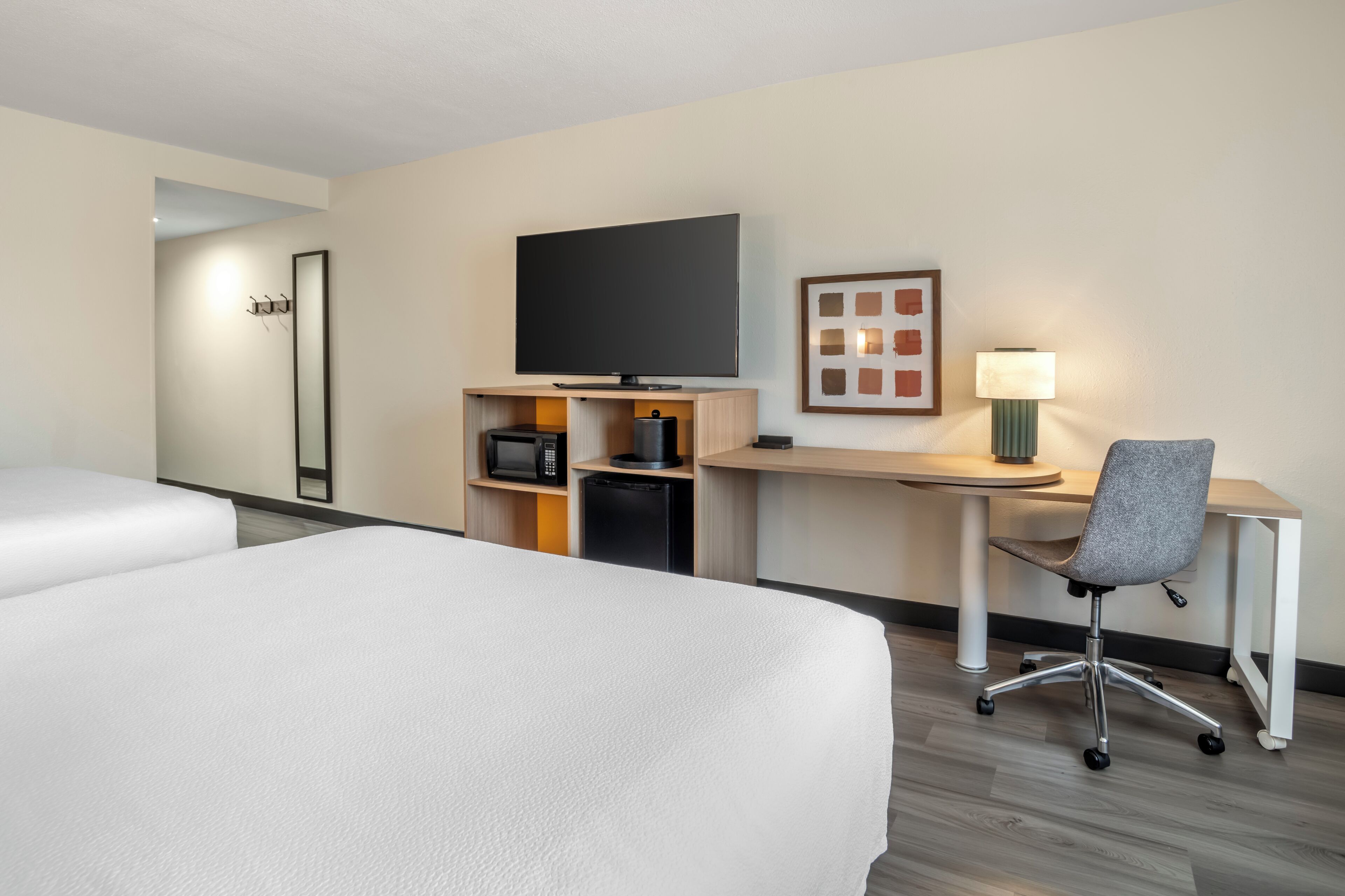 A hotel room with two beds, a desk with a chair, a television, a microwave, a refrigerator, and a framed picture on the wall.