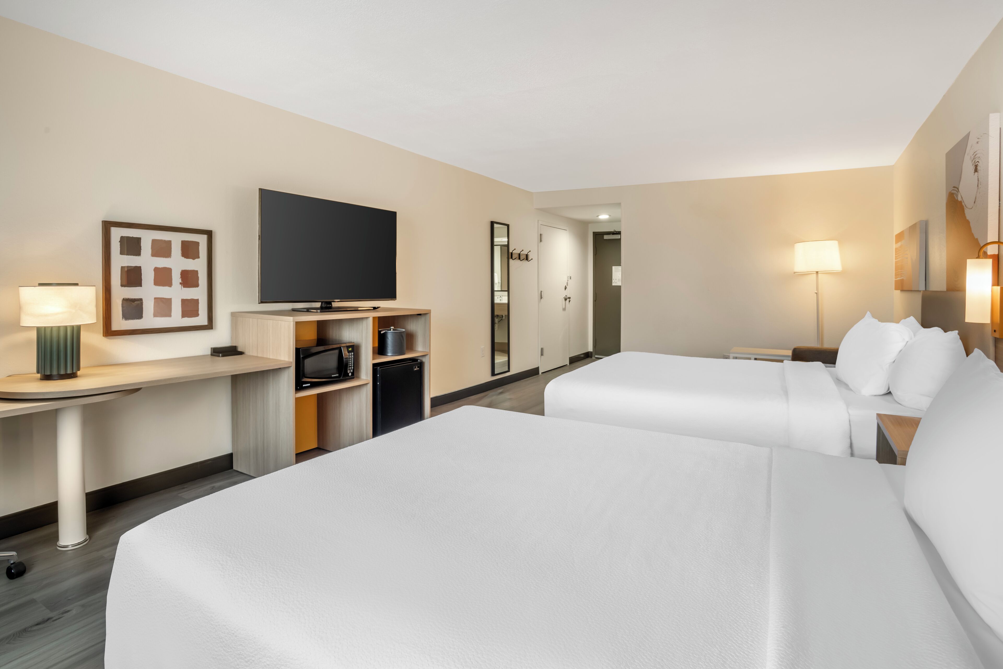 A hotel room with two beds, a TV, a desk with a lamp, a microwave, and a small refrigerator.