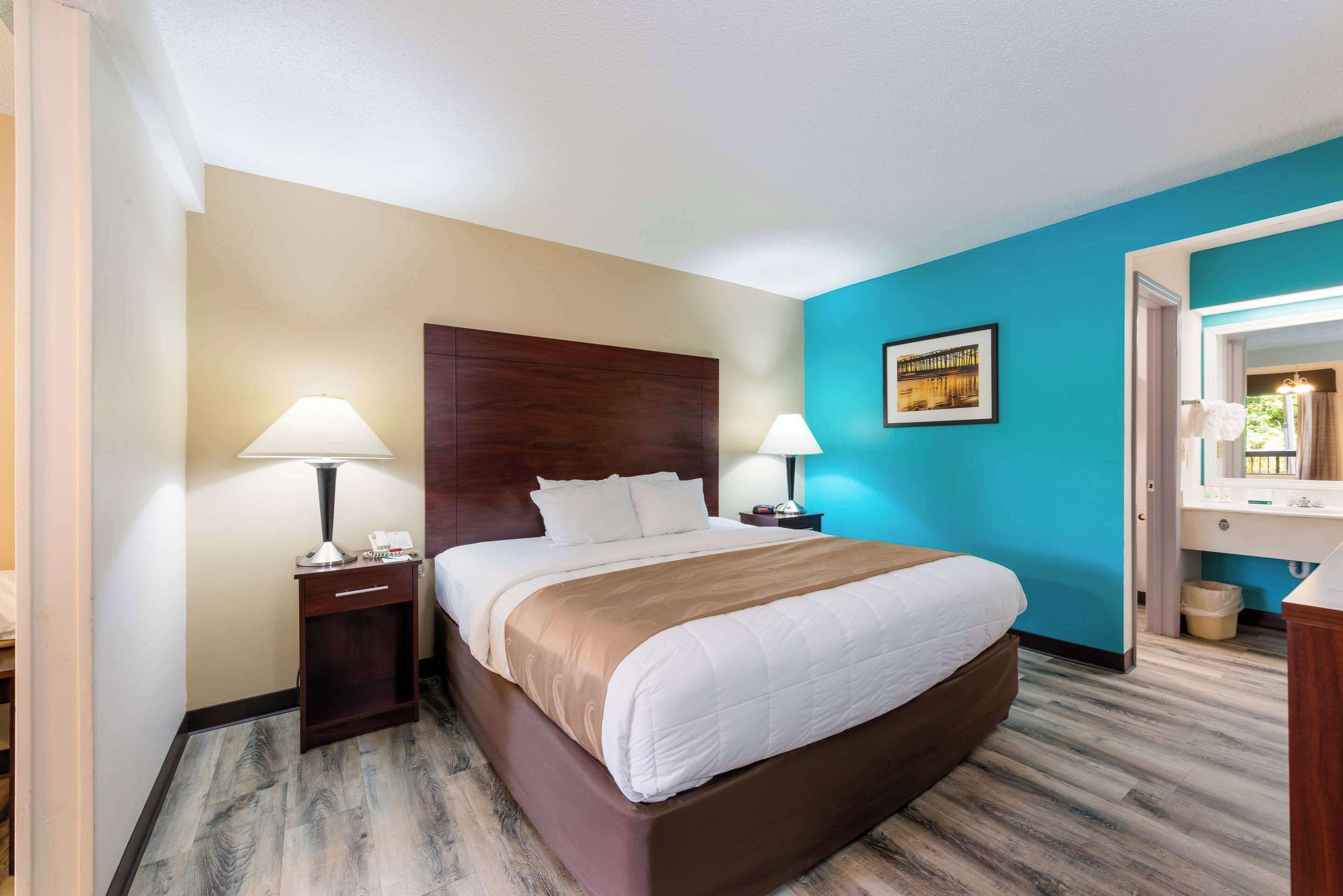 Une chambre d’hôtel avec un grand lit, deux tables de chevet avec des lampes, une table de nuit, une salle de bain avec un lavabo et un miroir, et un mur d’accent bleu.