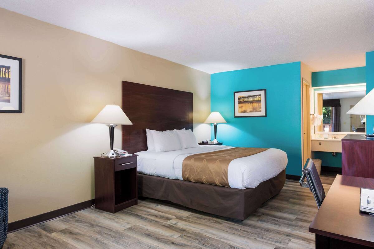 Ein Hotelzimmer mit einem großen Bett, zwei Nachttischen mit Lampen, einem Holz-Kopfteil, einem Schreibtisch mit Stuhl und einem Badebereich mit Spiegel und Waschbecken.
