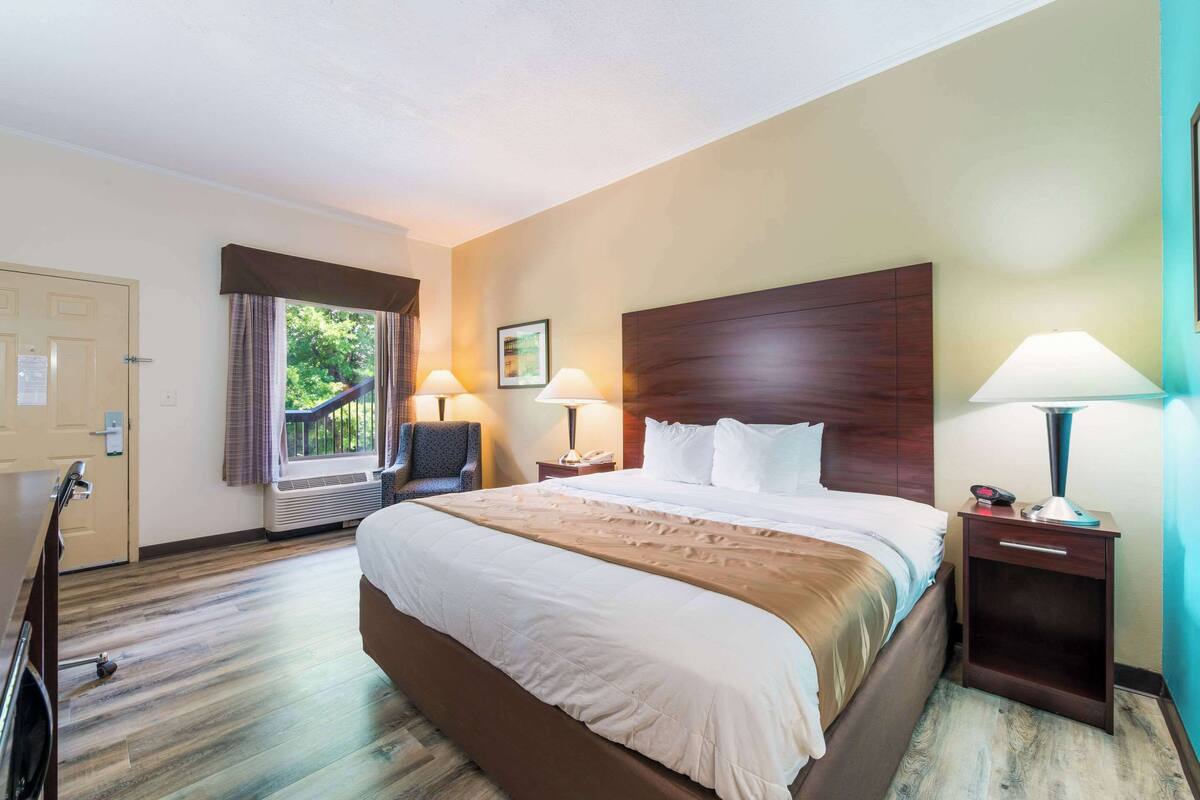 Ein Hotelzimmer mit einem großen Bett, zwei Nachttischen mit Lampen, einem Sessel, einer Tür und einem Fenster mit Vorhängen.