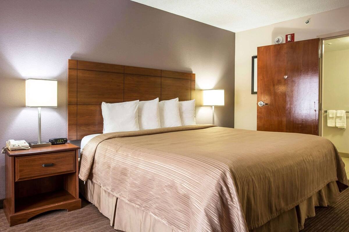 Una habitación de hotel con cama, lámparas de noche, mesita de noche, cabecero de madera y una puerta.