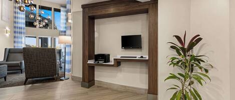 Un área de recepción moderna con un mostrador de madera, un televisor de pantalla plana, una planta en maceta y una zona de estar con una silla estampada y una lámpara.