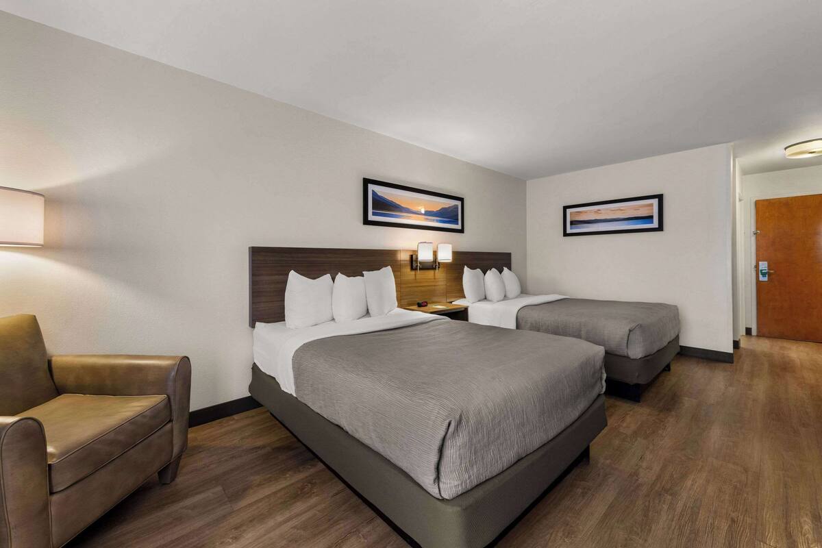 Una habitación de hotel con dos camas, una silla, una mesita de noche y dos cuadros en la pared.