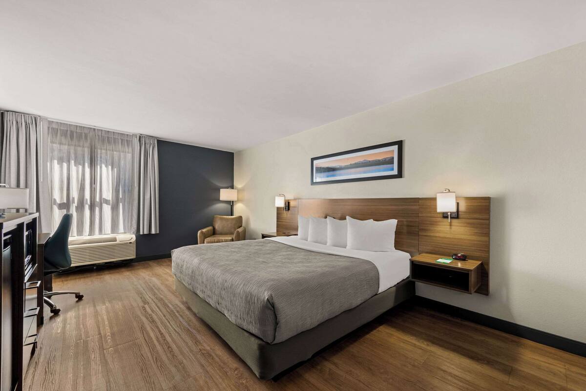 Habitación de hotel con una cama grande, un escritorio con silla, una lámpara de escritorio, una mesita de noche y una ventana con cortinas.