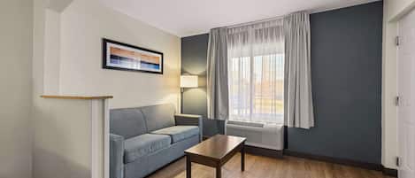 Een moderne woonkamer met een grijze bank, een houten salontafel, een ingelijste foto aan de muur, een staande lamp en een raam met gordijnen.