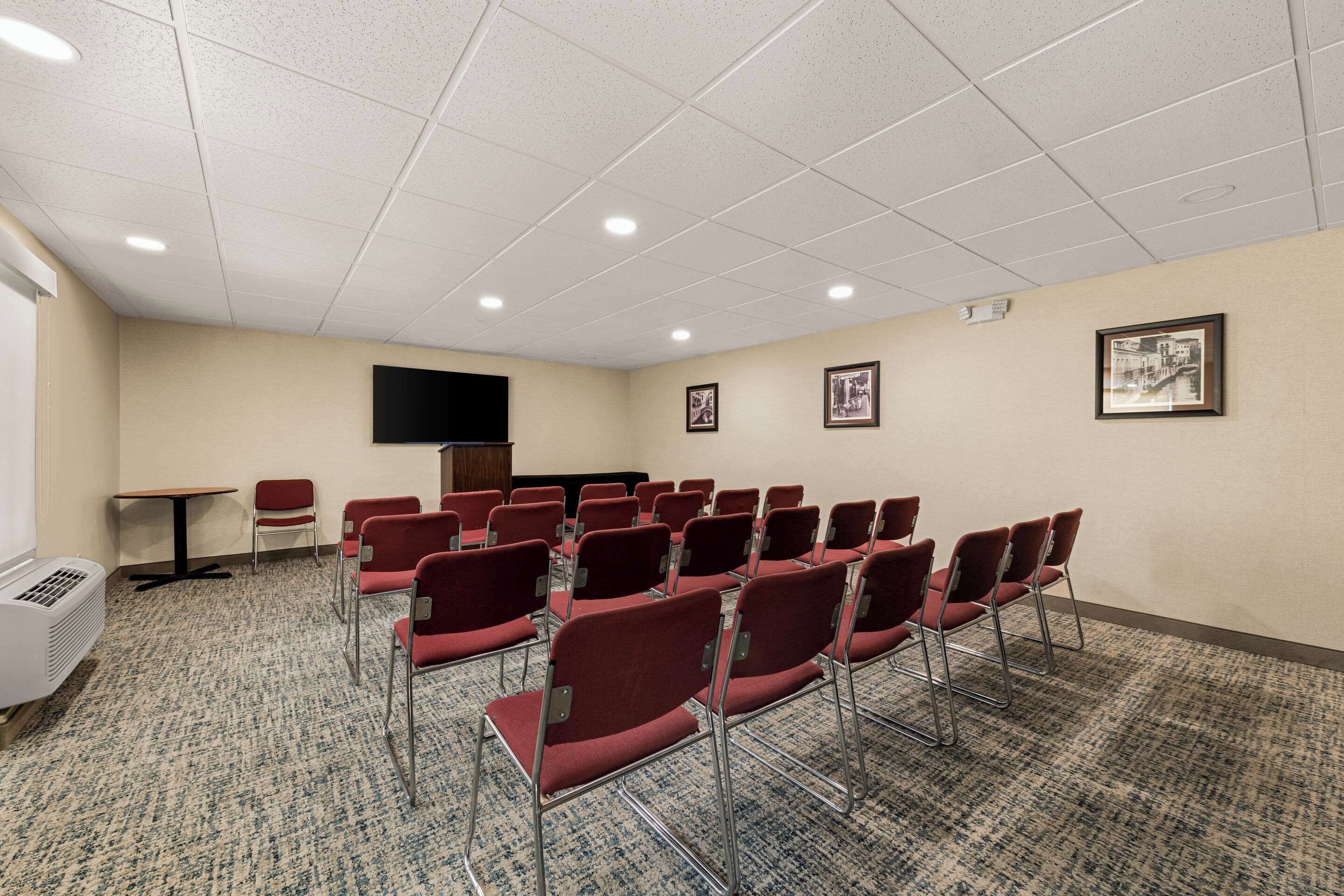 Un salón de conferencias con filas de sillas rojas, una pantalla de proyector y cuadros en la pared.