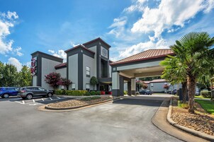 Un hotel moderno con vestíbulo cubierto, estacionamiento y una zona ajardinada con palmeras.