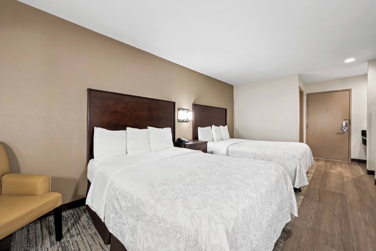 Quarto de hotel com duas camas, uma cadeira, uma escrivaninha e uma porta.