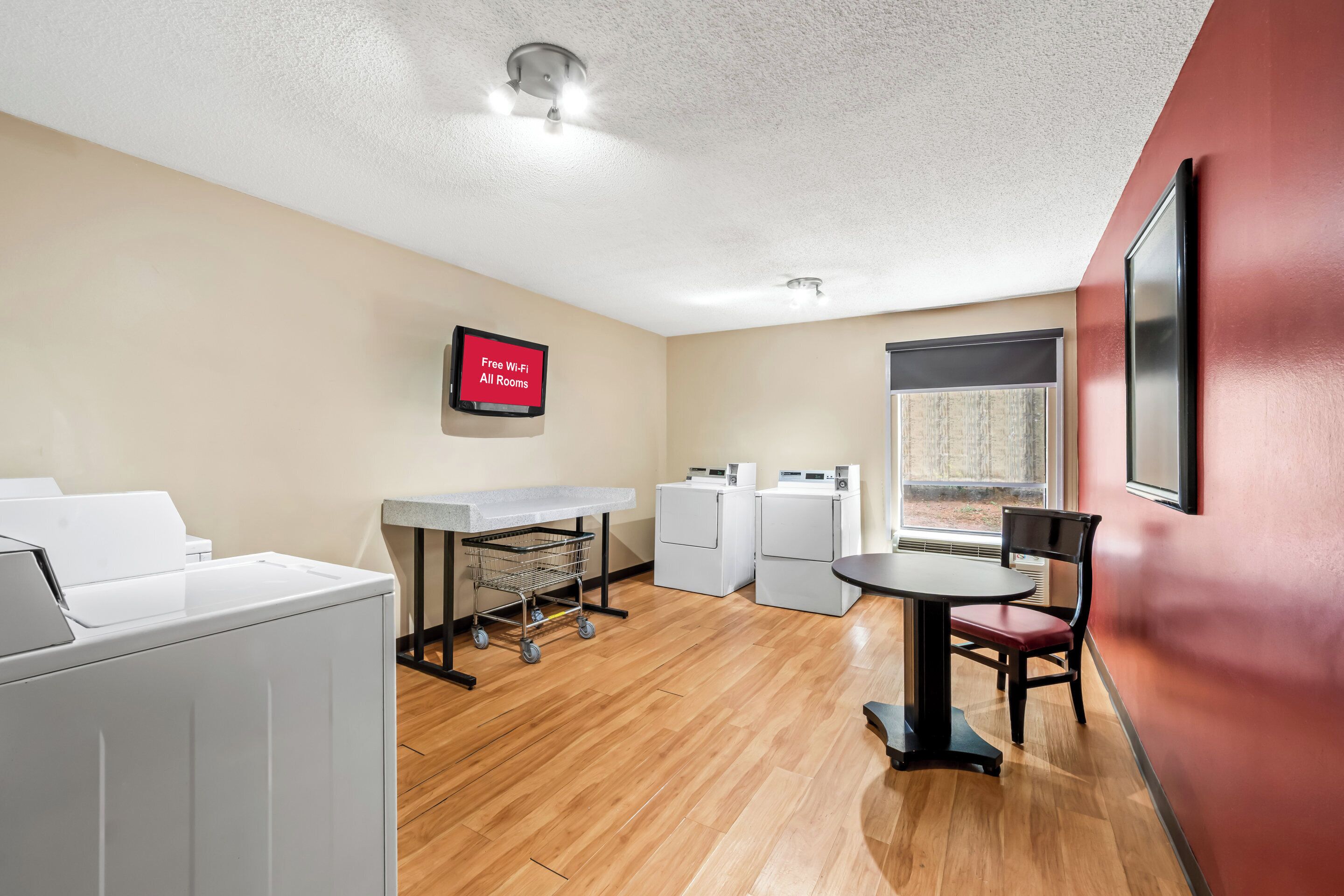 A hotel room with a kitchenette, a dining area, a laundry station, and a television displaying 'Free Wi-Fi All Rooms'.
