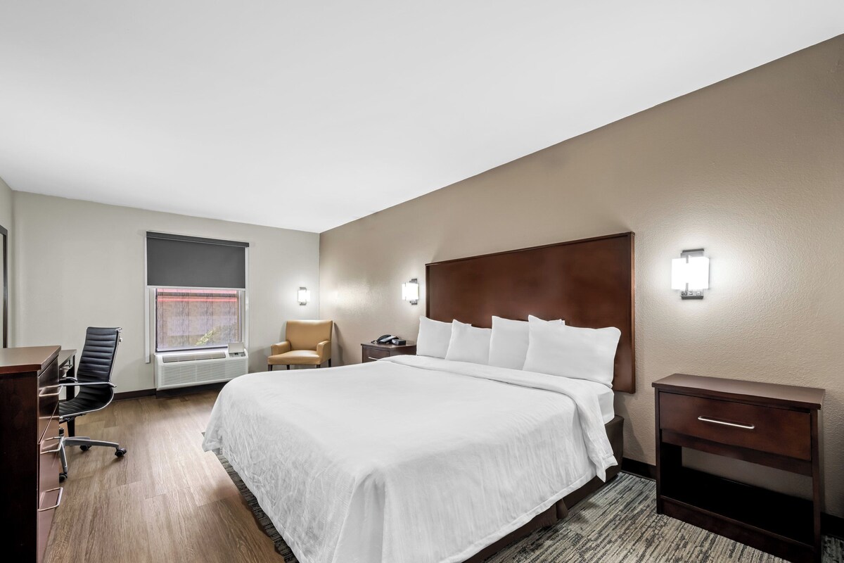 Une chambre d’hôtel avec un grand lit, un bureau, une chaise, une table de chevet et une fenêtre avec des stores.