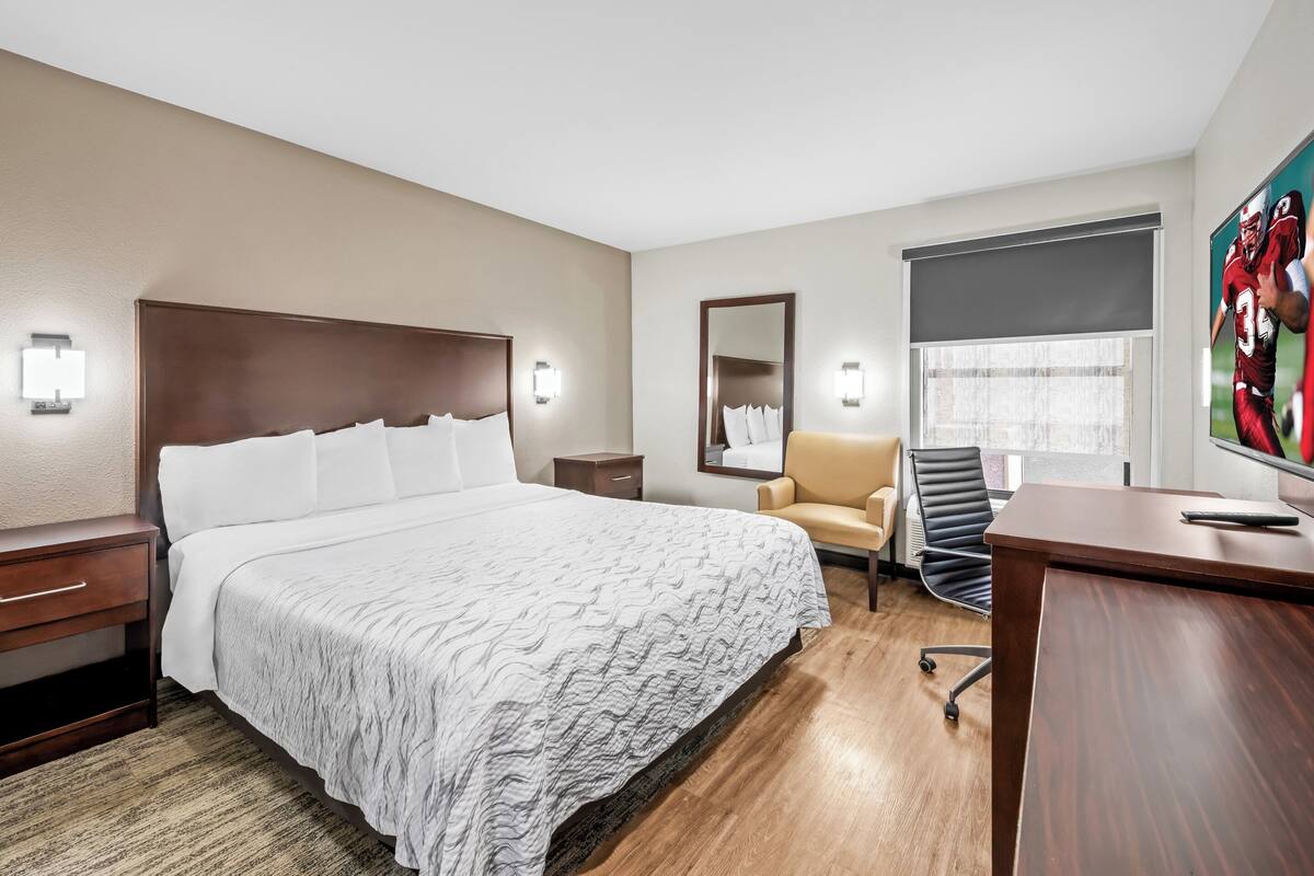 Quarto de hotel com uma cama grande, uma escrivaninha, uma cadeira, um espelho e uma janela com persianas.