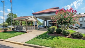 A building with a covered entrance, a parking lot, and a well-maintained lawn with flowering bushes.