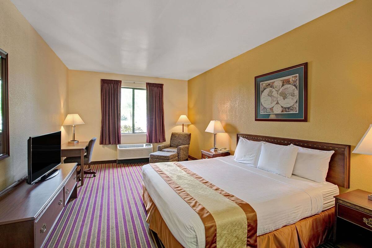 A hotel room with a large bed, a desk with a computer, two chairs, a TV, and a window with curtains.