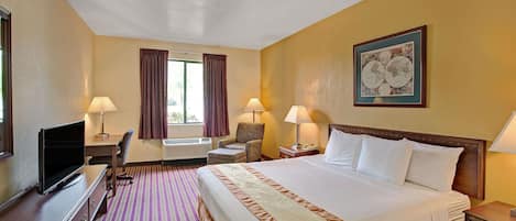A hotel room with a large bed, a desk with a computer, two chairs, a TV, and a window with curtains.