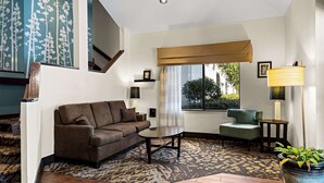 A living room with a brown sofa, a small round table, a green armchair, a large potted plant, and a patterned rug. - Sleep Inn Wilmington near University (Wilmington)
