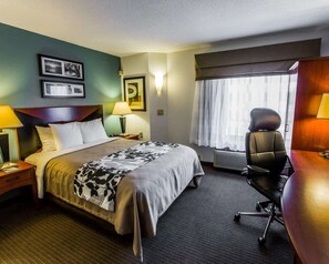A hotel room with a bed, a desk, a chair, a lamp, a picture frame, a window with curtains, and an air conditioning unit. - Sleep Inn Wilmington near University (Wilmington)