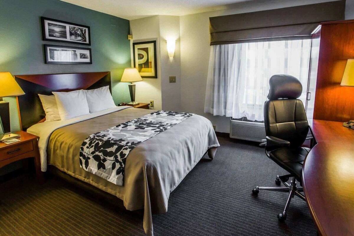 Ein Hotelzimmer mit Bett, Schreibtisch, Stuhl, Nachttisch, Lampe, Bilderrahmen, Fenster mit Vorhängen und Klimaanlage.