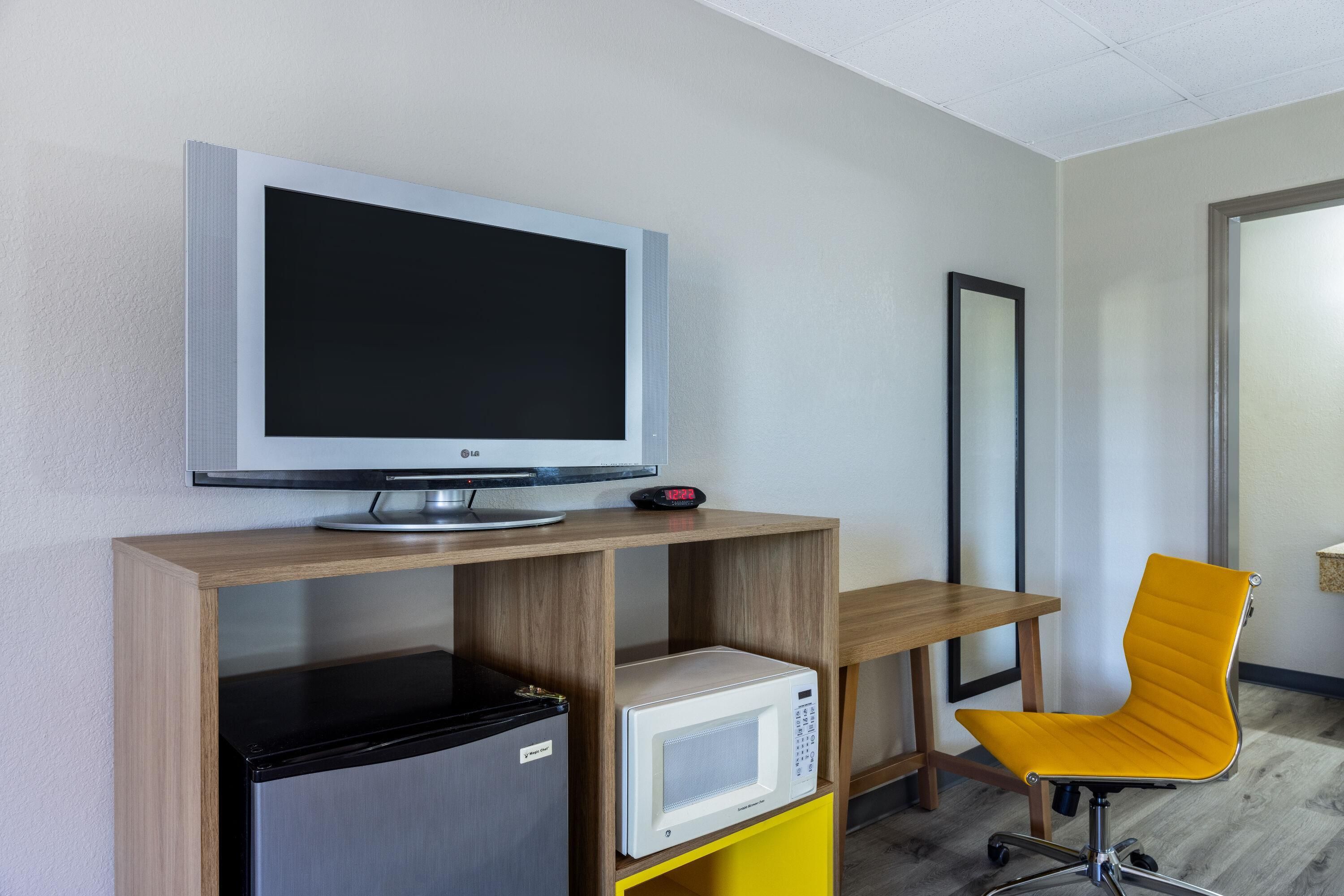 Una habitación moderna con televisión de pantalla plana sobre un soporte de madera, un refrigerador pequeño, un microondas y una silla amarilla.