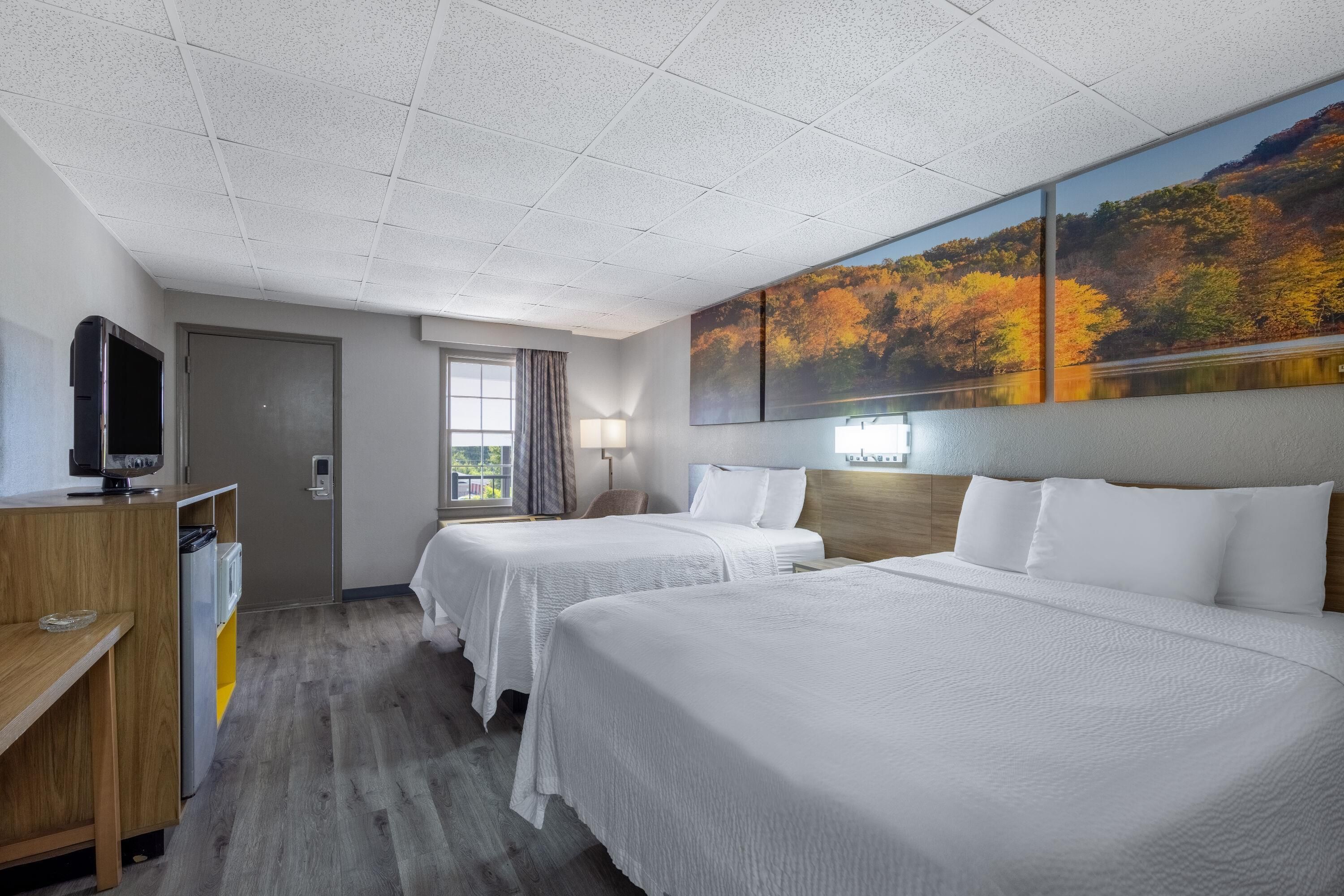 A hotel room with two beds, a television, a desk, and a painting of autumn trees.