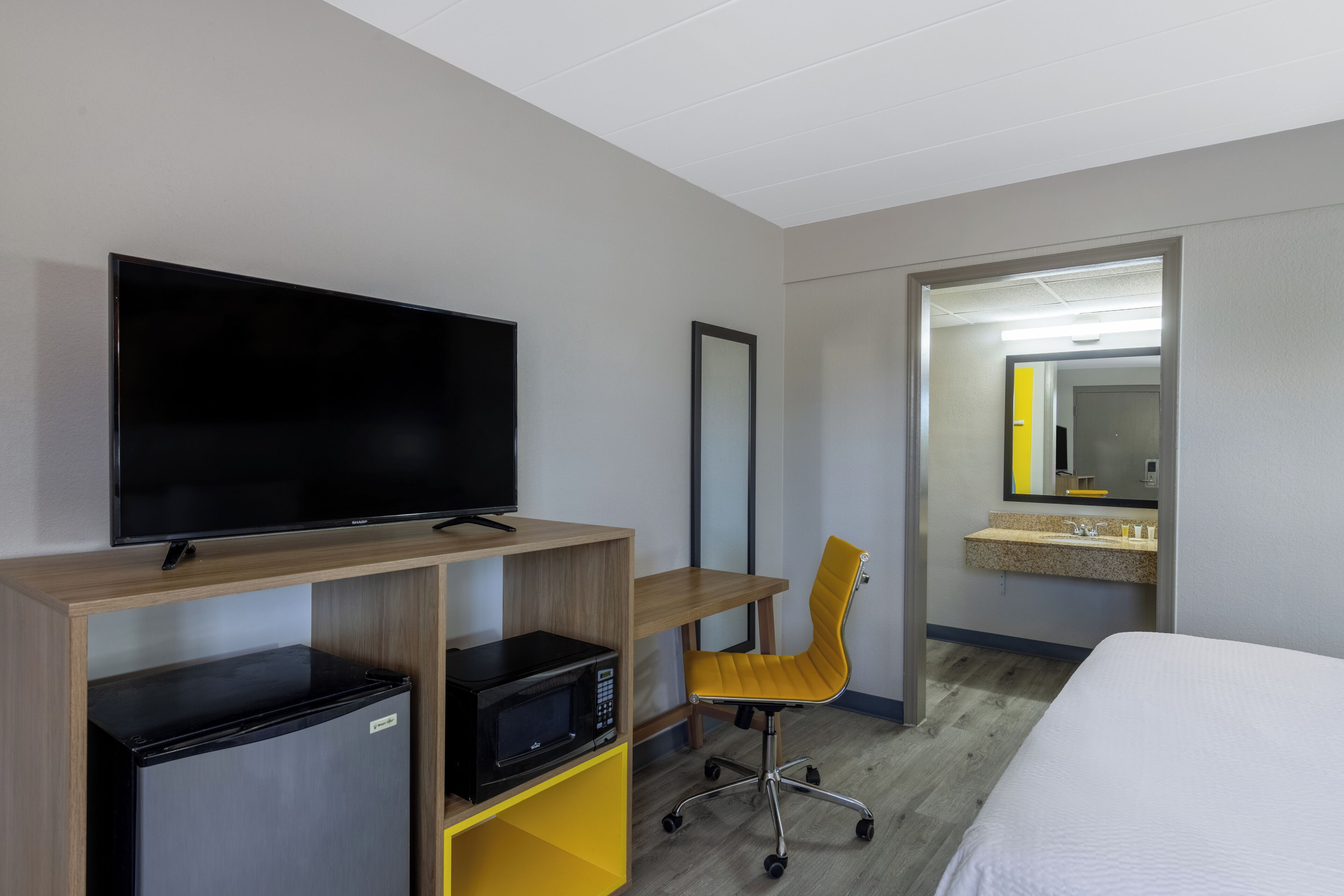 A hotel room with a flat-screen TV, a mini-fridge, a microwave, a yellow office chair, a bed, and a bathroom with a sink and mirror.