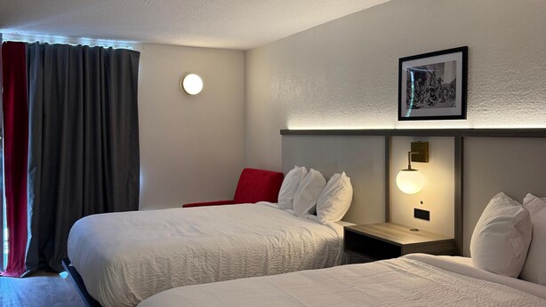 Ein Hotelzimmer mit zwei Betten, einem roten Sessel, einem Nachttisch mit Lampe und einem gerahmten Bild an der Wand.