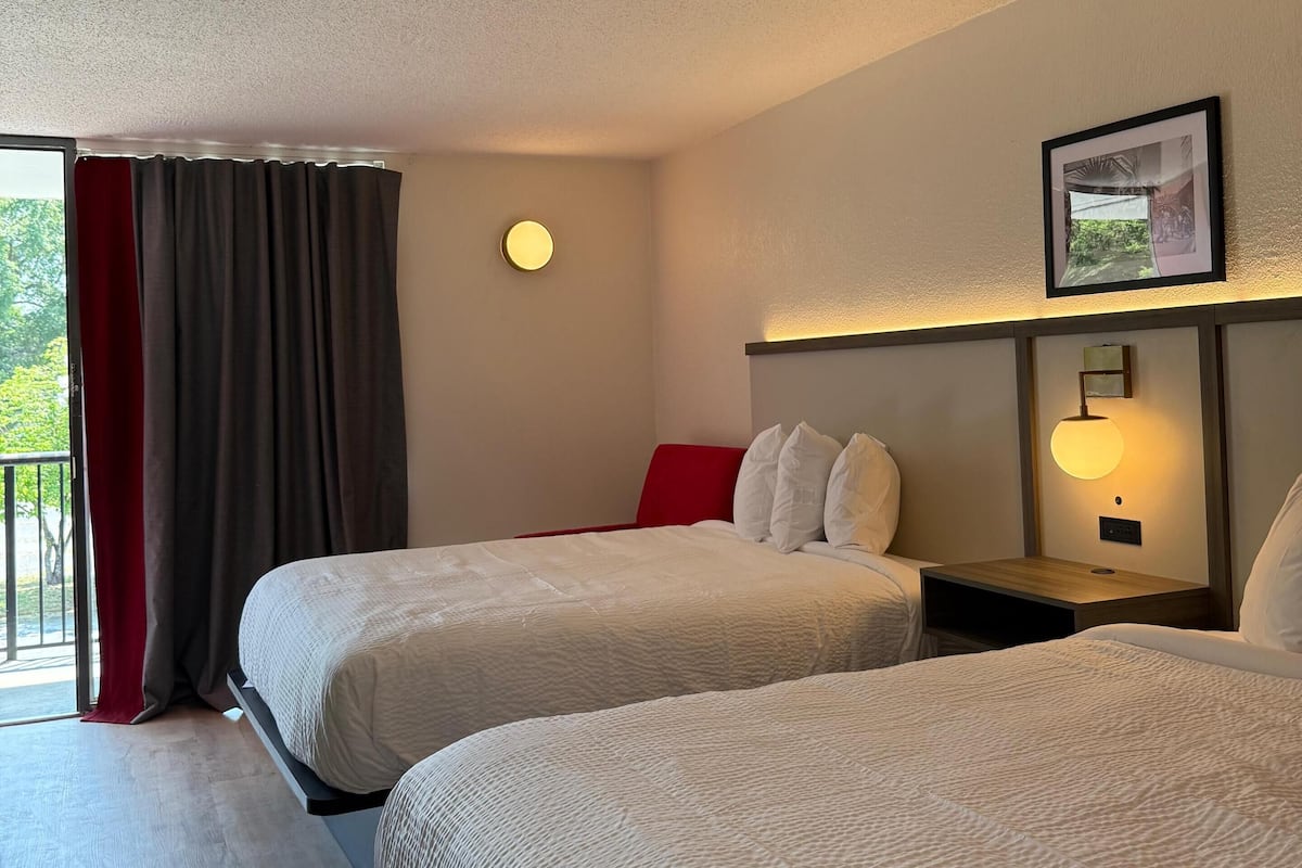 A hotel room with two beds, a red accent wall, a round wall light, and a framed picture on the wall.