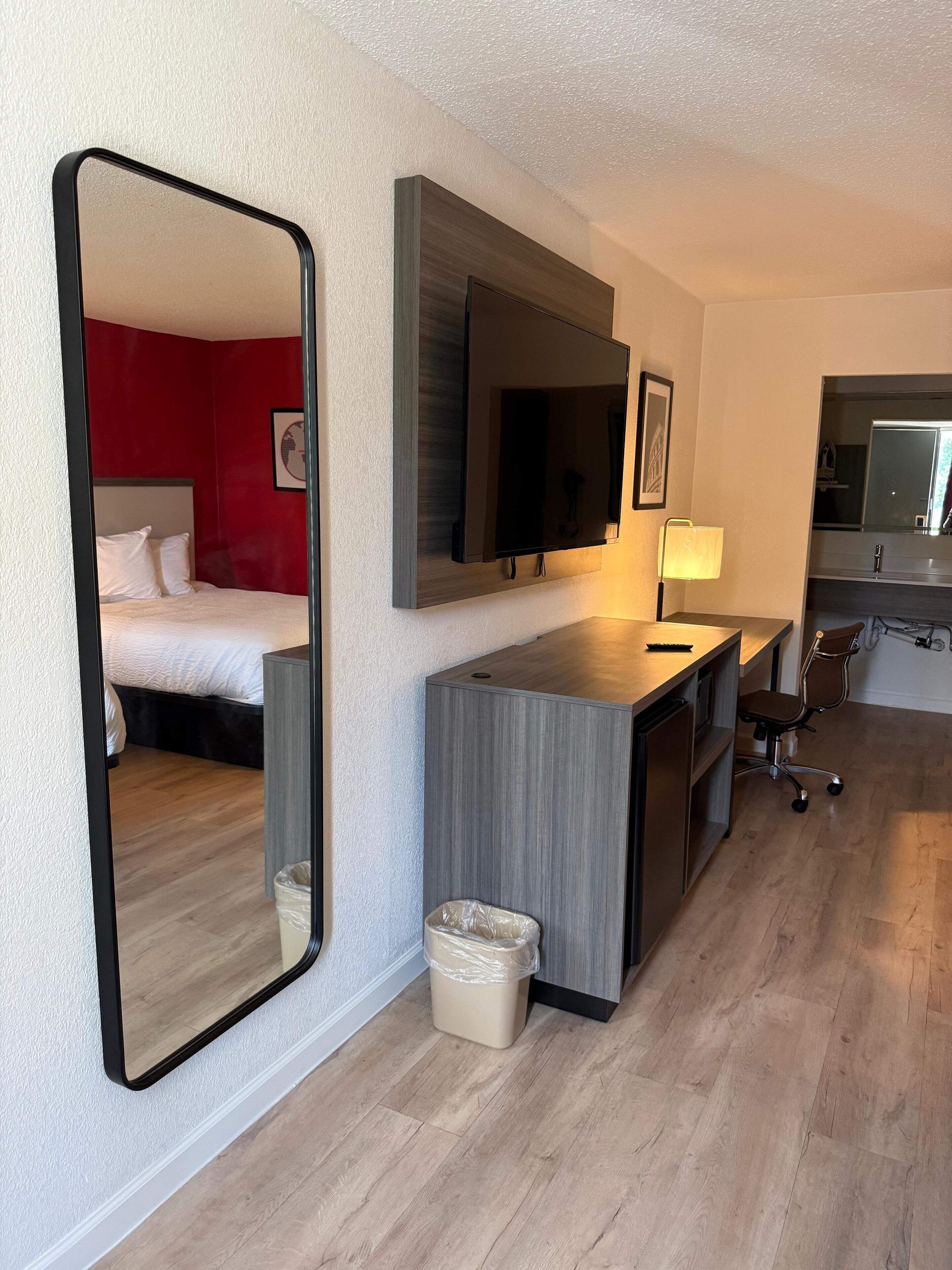 A hotel room with a bed, a desk, a chair, a mirror, and a trash bin.