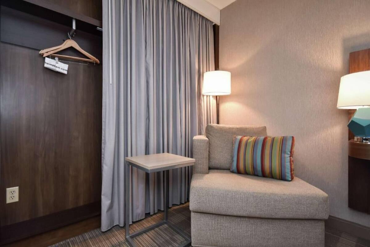 A hotel room with a grey sofa, a striped pillow, a small table, a hanging coat rack, and two wall-mounted lamps.