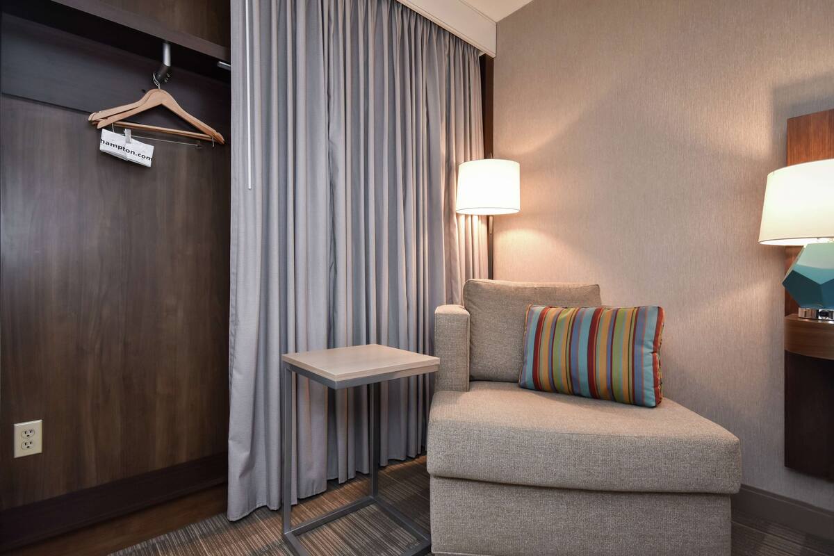 A hotel room with a grey sofa, a small side table, a lamp, and a hanging coat rack.