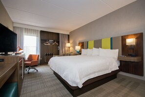 A hotel room with a large bed, a desk with a television, a chair, and a desk lamp.