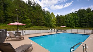 A rectangular swimming pool with a metal ladder on one side, surrounded by a black metal fence. There are two red and white umbrellas providing shade, and several lounge chairs and tables are arranged around the pool.