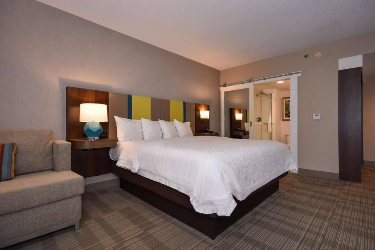 A hotel room with a bed, a nightstand, a lamp, a chair, and a mirror.