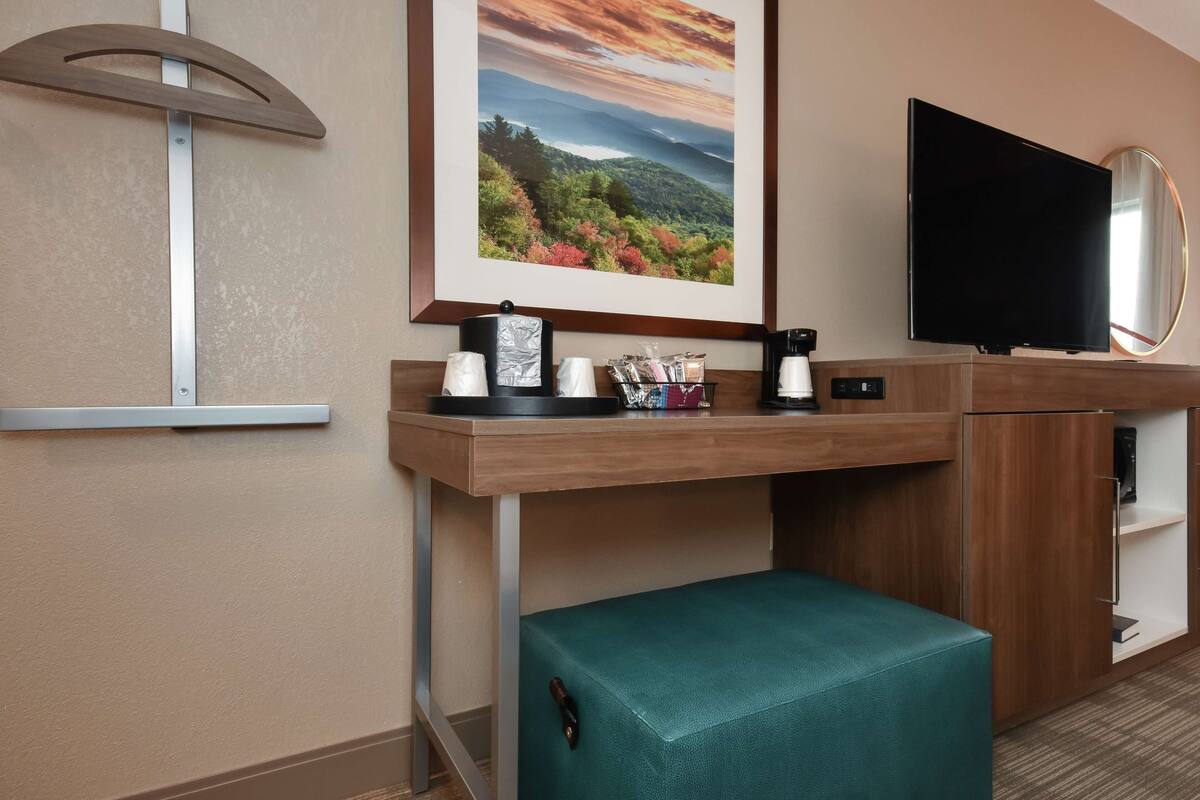 A hotel room with a desk, a flat-screen TV, a framed landscape photo, a small table with a cup and a tissue box, and a green cushioned bench.
