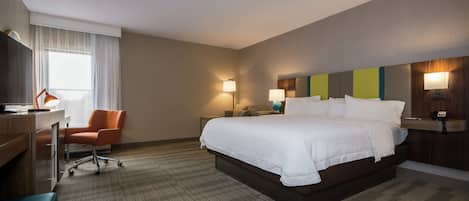 Une chambre d’hôtel avec un grand lit, un bureau, une chaise orange et une fenêtre avec des rideaux blancs.