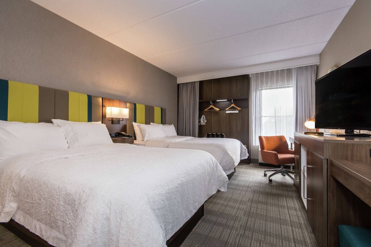 A hotel room with two large beds, a desk with a television, a chair, and a desk lamp.