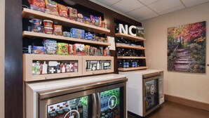 A snack display with various chips and snacks, a refrigerator with beverages, and a painting of a forest on the wall. - Hampton Inn Brevard (Pisgah Forest)