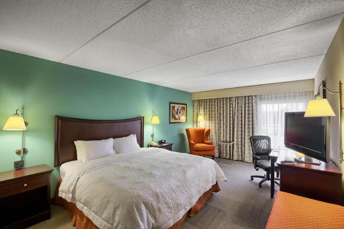 Habitación de hotel con cama, escritorio con computadora, silla, televisor y ventana con cortinas.