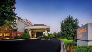Entrada de un hotel Hampton Inn con un paseo techado, jardín ajardinado y cielo despejado.