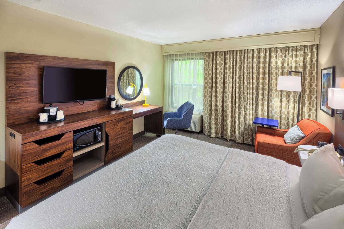 Habitación de hotel con una cama grande, un televisor de pantalla plana montado en un mueble de madera, un escritorio con una silla, una mesita con una lámpara y una ventana con cortinas.