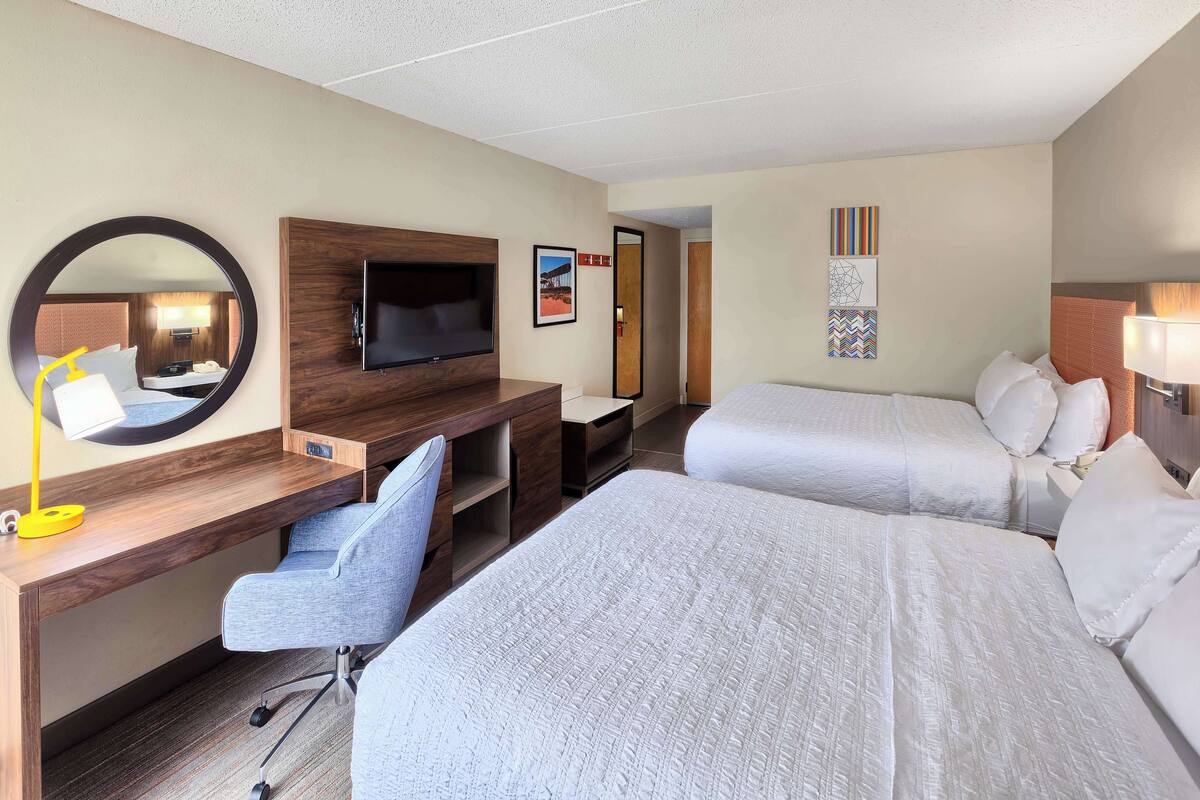 Habitación de hotel con dos camas, un escritorio, una silla, un televisor y obras de arte en las paredes.
