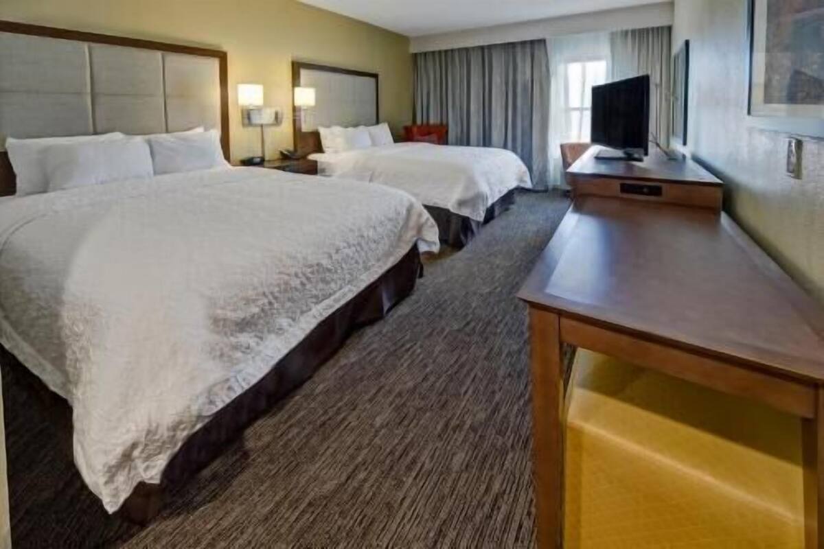 A hotel room with two beds, a desk, a TV, and a window with curtains.
