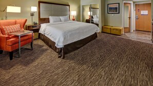 A hotel room with a large bed, a desk, an orange armchair, a mirror, and a door. - Hampton Inn by Hilton Concord/Kannapolis (Concord)