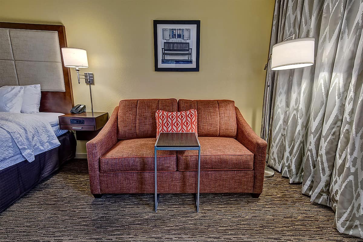 A hotel room with a bed, a sofa, a small table, a lamp, a framed picture on the wall, and a curtain.