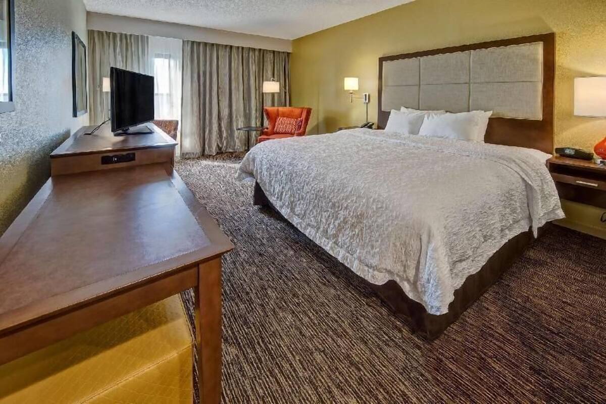 A hotel room with a large bed, a desk with a television, a chair, and a nightstand with a lamp.
