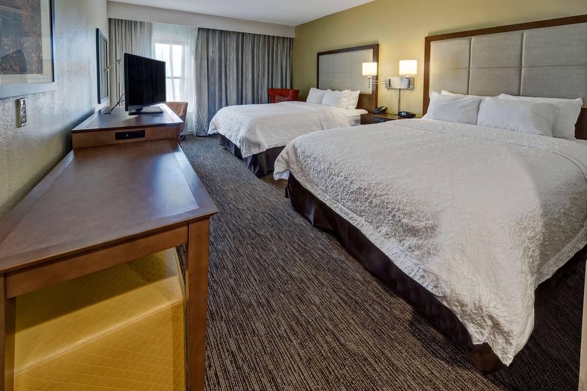 A hotel room with two beds, a desk, a television, and a large window with curtains.
