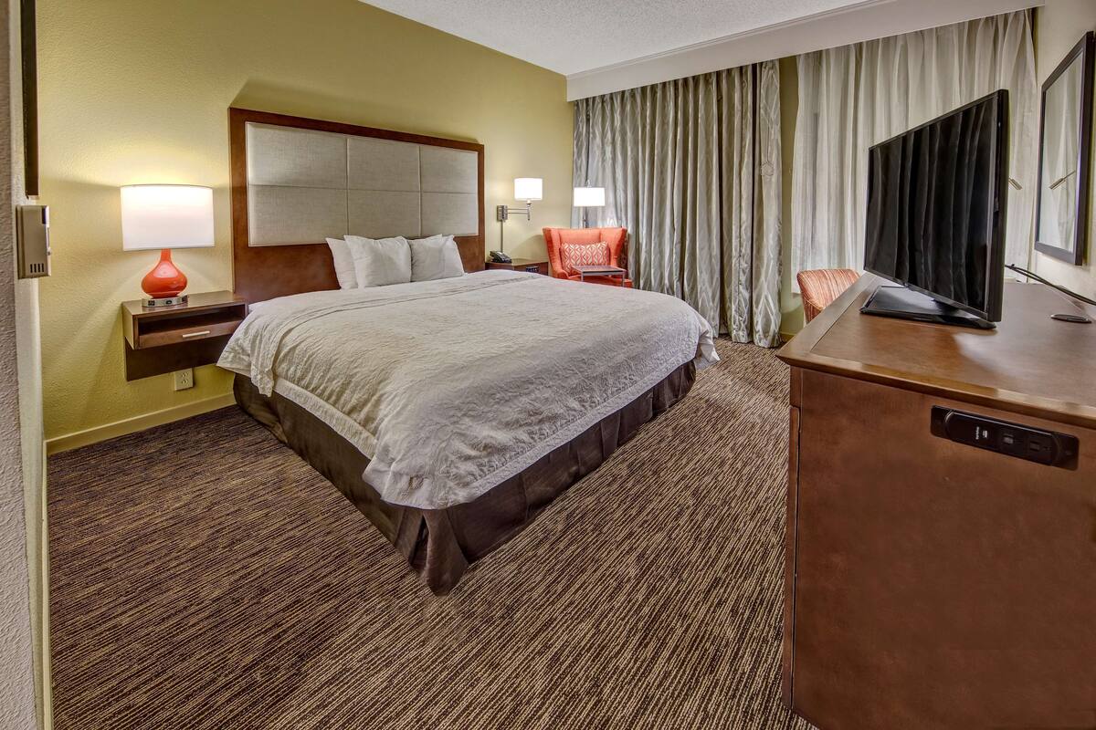 A hotel room with a large bed, a desk with a television, a chair, and two lamps.